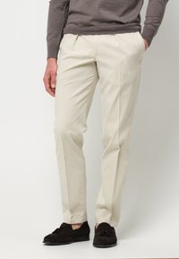 Beige maßgeschneiderte Hose mit flachem Bund, aus glattem Stoff. Kombiniert mit dunkelbraunen Wildlederschuhen mit Quastendetail.