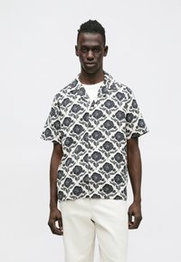 Koszula z krótkim rękawem z czarnym kwiatowym wzorem na białym tle, o luźnym kroju i kołnierzykiem z wycięciem. Zestawiona z jasnymi spodniami.