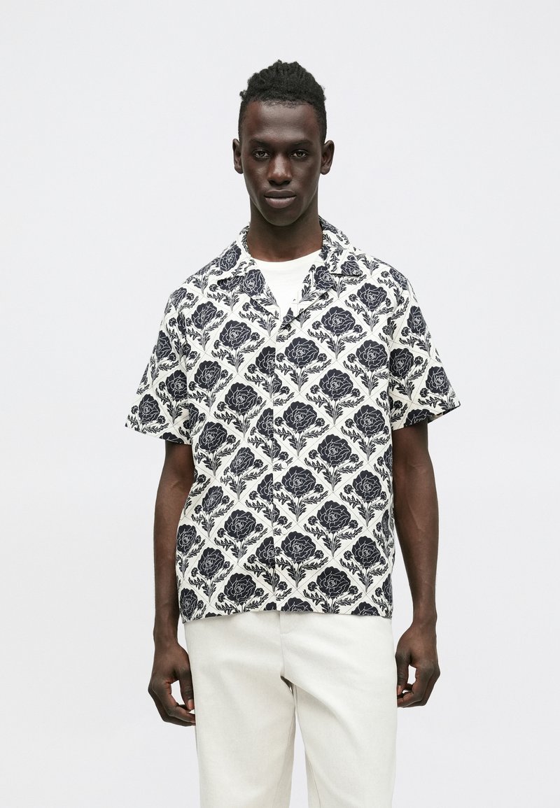 Koszula z krótkim rękawem z czarnym kwiatowym wzorem na białym tle, o luźnym kroju i kołnierzykiem z wycięciem. Zestawiona z jasnymi spodniami.