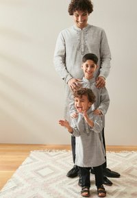 Tre individer bär matchande ljusgrå, broderade tunikor med knappar och står på en mönstrad matta, med glada uttryck.