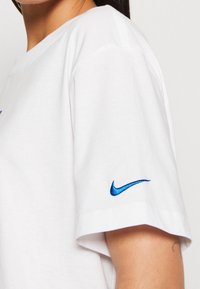 Vit bomull T-shirt med korta ärmar, med en blå broderad Nike-logotyp på vänster ärm. Slätt tyg och avslappnad passform.