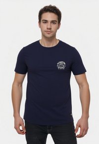 Camiseta de color azul marino hecha de algodón, con cuello redondo y mangas cortas. Incluye texto del logo en blanco en el área del lado izquierdo del pecho.