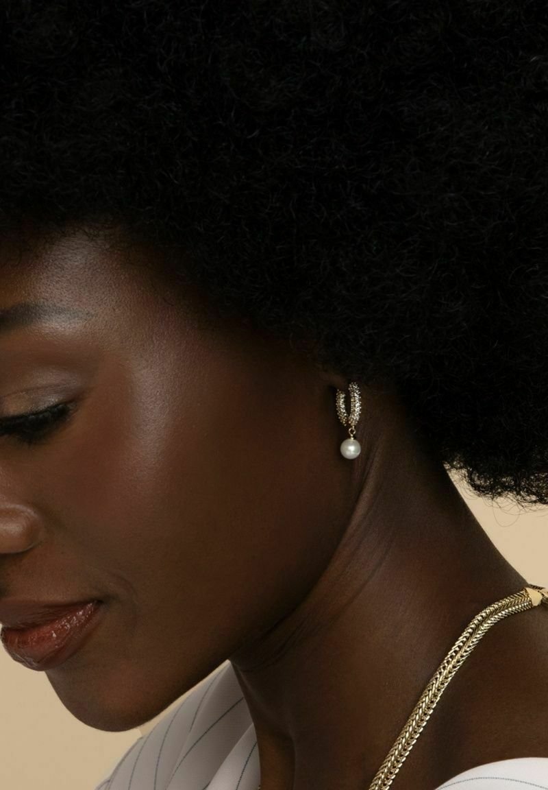 Guldörhängen med texturerad design, som har en hängande pärla. Kort, delvis profilbild med naturligt, lockigt svart hår synligt.