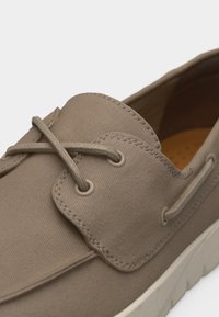 Mocassin décontracté beige avec détails cousus, lacets croisés passant par des œillets métalliques, et une semelle en caoutchouc blanche.