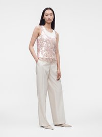 Femme portant un haut sans manches blanc sous un débardeur rose à sequins, associé à un pantalon large crème et des chaussures pointues, debout avec une main dans la poche.