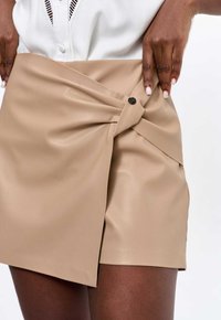 Beige imitatieleren wikkelrok met een asymmetrisch ontwerp, een voorkant met twist en een zwarte knoopsluiting aan de zijkant. Glad textuur.