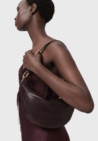 Bruine leren halve maantasje met een gladde textuur, gouden hardware accenten, gedragen aan een schouderriem, gecombineerd met een donker bordeauxrode jurk.