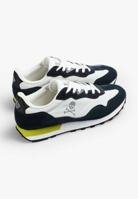 Sneaker bianco e navy con tomaia in suede e tela, caratterizzato da una grafica di teschio, suola in gomma nera e accentuazione gialla sul tallone.