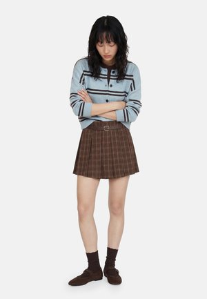 Ung kvinde med krydsede arme iført en lyseblå stribet cardigan, brun ternet nederdel, mørke sokker og brune sko, stående mod hvid baggrund.