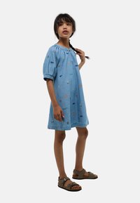 Robe en denim avec des manches bouffantes courtes, de couleur bleue, arborant des motifs brodés colorés de la nature et d'éléments extérieurs. Sandales marron.