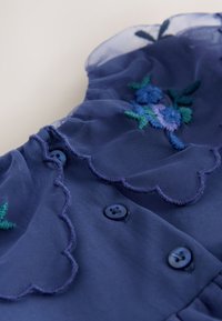 Vestido azul marino con capas transparentes, bordes festoneados, diseños florales bordados en azul y verde, y botones azul oscuro para cierre.