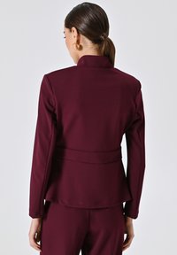 Une femme aux cheveux bruns attachés en chignon bas porte un blazer ajusté bordeaux foncé et un pantalon assorti, vue de dos sur fond uni.