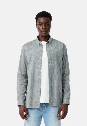 Hombre con camisa abotonada de patrón gris claro sobre una camiseta blanca y vaqueros azul claro, de pie frente a un fondo blanco liso.