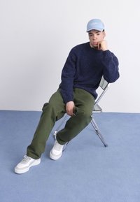 Suéter de punto navy, pantalones verde oliva y zapatillas blancas. La gorra que lleva puesta es de color azul claro. El sujeto está sentado en una silla de metal sobre un suelo azul.