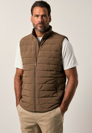 Brown puffer vest with a high collar, horizontal stitching, and zippered front. Worn over a white shirt, paired with light-colored pants.