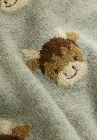 A zöld szövet texturált kötött mintázattal rendelkezik, amelyen mosolygó barna tehénfejek láthatók, krémszínű fülekkel és részletekkel kiemelve.