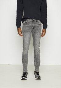 Jeans skinny gris oscuro con un lavado descolorido, cinco bolsillos y detalles de costura sutiles, combinados con una camiseta negra de manga larga y zapatillas deportivas.