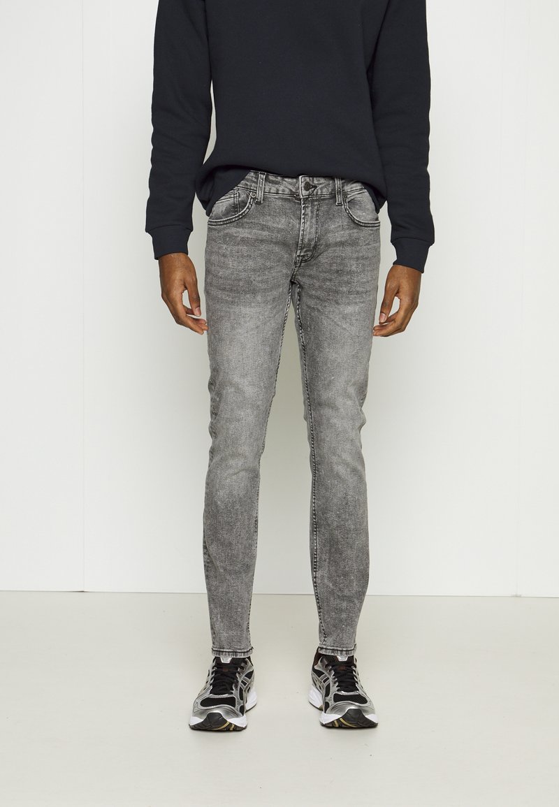 Jeans skinny gris oscuro con un lavado descolorido, cinco bolsillos y detalles de costura sutiles, combinados con una camiseta negra de manga larga y zapatillas deportivas.