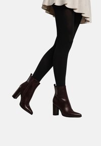 Bordeauxrode leren enkellaarzen met een spits toelopende neus en een stevige hak, voorzien van een zijrits en een gladde textuur, gedragen met zwarte panty's en een beige rok.