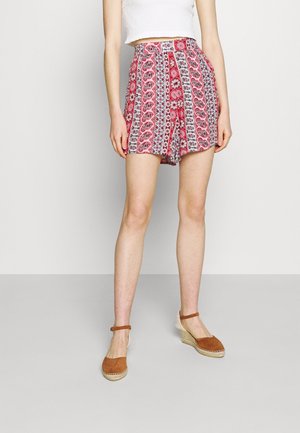 Un short taille haute rouge et blanc à motifs, avec des plis, associé à des chaussures espadrilles marron. Le tissu présente des motifs floraux et paisley.
