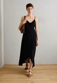 Black sleeveless dress with a draped neckline and asymmetrical hem, paired with patterned footwear and a brown shoulder bag.