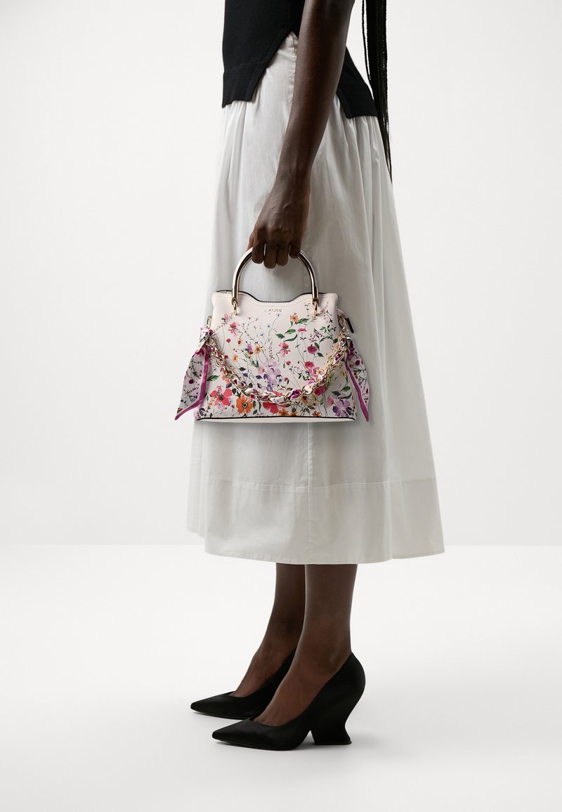 Femme en jupe blanche et haut noir tient un petit sac blanc avec un imprimé floral coloré et un détail de chaîne dorée.