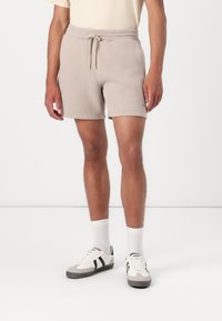 Pantalones cortos de algodón color beige claro con cintura elástica y cordón ajustable. Con bolsillos laterales y dobladillos enrollados. Combinados con calcetines blancos y zapatillas.