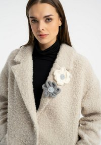 Beżowy wełniany płaszcz o fakturowanym wykończeniu, z dwoma broszkami w kształcie kwiatów w odcieniach szarości i bieli z okrągłymi metalicznymi akcentami. Czarny golf pod spodem.