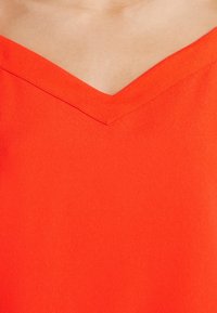 Haut rouge à col en V fabriqué en tissu lisse, avec un décolleté en forme de V accentué et des bords sans coutures. Pas de motifs ni d'accents visibles.