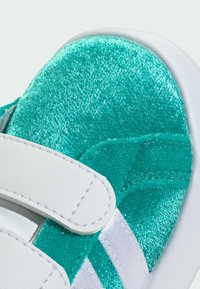 Tyrkysové tenisky s plyšovým zvrškom, bielymi nastaviteľnými pásikmi a metalickými doplnkami. Hladká podrážka s textúrovanými akcentmi.