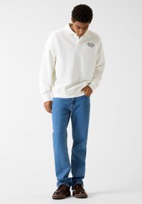 White long-sleeve polo with a collar, featuring blue embroidery. Paired with light blue straight-leg jeans and brown lace-up shoes.
