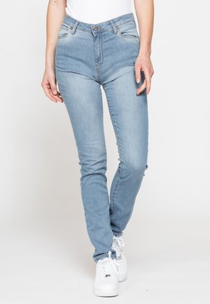 Jeans slim fit azzurro chiaro indossati con sneakers bianche e una canotta bianca, mostrati su una persona in piedi davanti a uno sfondo chiaro e semplice.