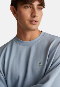 Sudadera azul claro con cuello redondo, hecha de tela suave. Presenta un pequeño logo bordado en verde en la zona del pecho izquierdo.