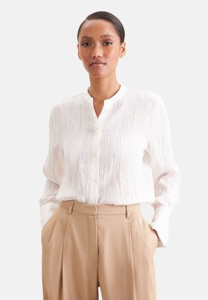 Femme portant un chemisier blanc texturé boutonné, rentré dans un pantalon beige plissé taille haute, les mains dans les poches.