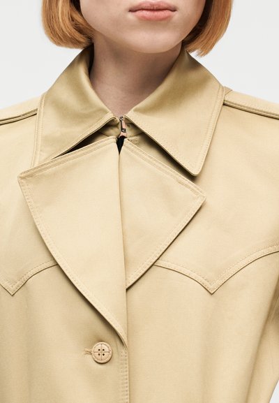 Γυναίκα που φοράει ένα μπεζ καμπαρντίνα με φαρδιές πέτσες, ένα κουμπί και φερμουάρ κάτω από το γιακά, κομμένο στο λαιμό και τους ώμους.