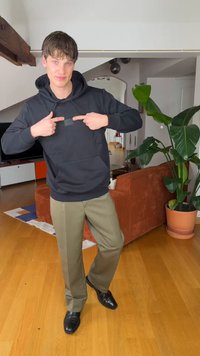 Ung mand iført sort hættetrøje og beige bukser peger mod brystet, stående på trægulv nær orange sofa og stor potteplante i stuen.