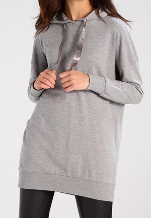 Woman wearing a grey hooded tunic sweatshirt with satin drawstrings and black leggings against a plain background.