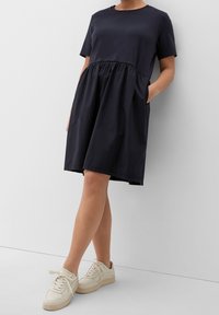 Robe en mélange de coton bleu marine avec un haut ajusté, une taille froncée et des poches latérales. Associée à des baskets beige clair.
