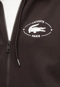 Veste noire zippée avec logo Lacoste blanc représentant un crocodile et le texte "Lacoste Paris" sur la poitrine près de la fermeture éclair.