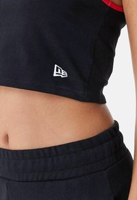 Top corto nero con dettagli rossi e una toppa bianca con logo; abbinato a pantaloni neri con cordino e vita strutturata.