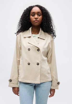 Femeie cu păr creț, purtând o jachetă bej cu două rânduri de nasturi și jeansi albastru deschis, stând pe un fundal alb simplu.