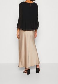 Schwarze, plissierte Bluse mit einem Ausschnitt auf der Rückseite, kombiniert mit einem glatten, beigefarbenen Satin-Maxikleid. Das Outfit wird durch schwarze Sandalen mit Blockabsätzen ergänzt.