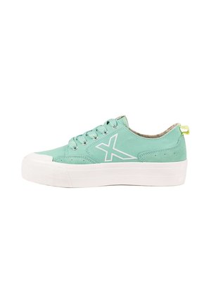 Zapatilla baja turquesa con suela blanca gruesa, logo blanco "X" en el lateral, cordones turquesa y lengüeta amarilla en el talón.