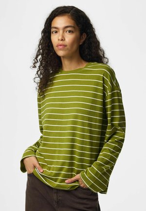 Femme aux longs cheveux bouclés portant une chemise verte ample à manches longues avec des rayures blanches horizontales et un pantalon foncé, les mains dans les poches.