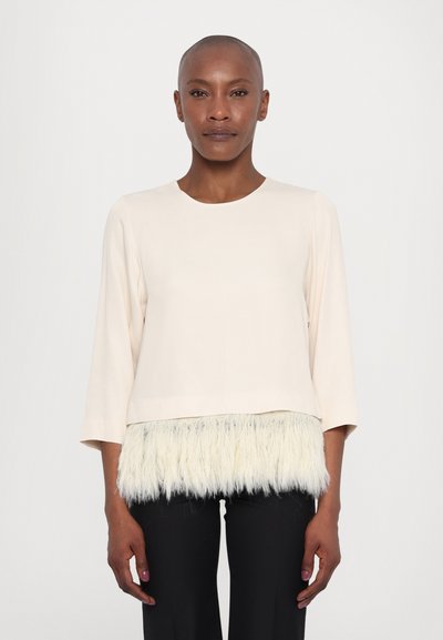 Light beige top with three-quarter sleeves, featuring a fluffy white fur trim at the hem. The fabric appears smooth and lightweight.