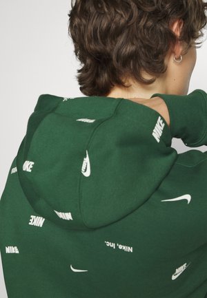 Grøn hættebluse lavet af blødt stof, med spredte hvide Nike-logoer og en afslappet pasform. Detaljeret syning langs kanterne.