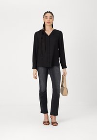 Blouse noire à manches longues avec un décolleté en V, associée à un jean sombre. Le mannequin tient un sac à main beige et porte des sandales à brides marron.
