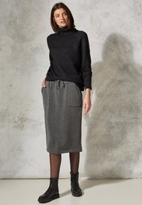 Schwarzer Rollkragenpullover mit gerippter Textur, kombiniert mit einem grauen knielangen Rock mit zwei Fronttaschen. Schwarze Schnürstiefeletten.