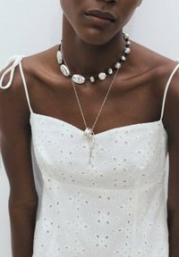 Abito bianco ricamato con scollatura quadrata e spalline sottili. Gioielli argentati includono una collana grossa e un pendente a strati.