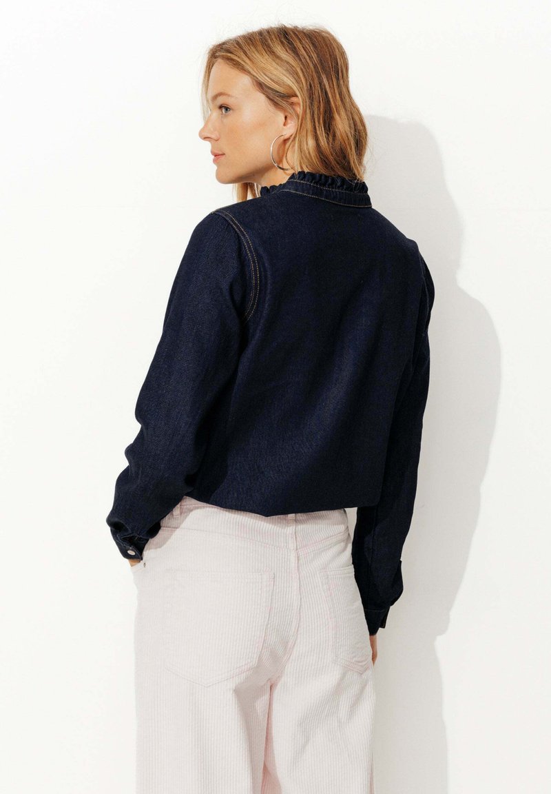 Chemise en denim foncé avec un col volanté, présentant des manches longues et des poignets boutonnés, associée à un pantalon en velours côtelé rose clair.